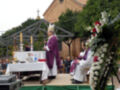 Una imagen de una ceremonia religiosa al aire libre, con un sacerdote en vestimenta roja y blanca frente a una mesa cubierta con mantel blanco. Hay flores blancas y rojas en un pedestal a la derecha, y una estructura de madera con farolillos encendidos en el fondo.