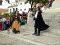Dos personajes históricos, una mujer con vestimenta medieval y otro en traje negro, caminan por las escaleras de un edificio blanco. La escena está rodeada de espectadores observando la actuación, con un ambiente que sugiere una representación o evento histórico.