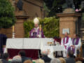 El obispo, vestido de morado y con un manto blanco, se encuentra en el centro de la imagen, frente a una mesa cubierta con mantel blanco y rojo. A su lado, dos sacerdotes vestidos de blanco con manto morado están asistiendo a la ceremonia. En el fondo, se pueden ver plantas y una estructura de ladrillo.