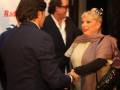 Una mujer con cabello rubio y una bufanda beige se está estrechando la mano de un hombre en un evento formal.