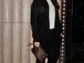 A una mujer con un look elegante y casual. Lleva un abrigo negro largo, una camiseta blanca con cuello redondo y pantalones negros ajustados. Complementa su look con un clutch de piel estampada y sandalias negras. La背景包括一个米色的窗帘和一扇装饰有圆点图案的大窗户。