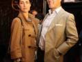 A una pareja en un evento formal. La mujer lleva abrigo de lana marrón y el hombre está con una chaqueta beige. Ambos están sonrientes, posando para la foto en un fondo con estrellas y luces.