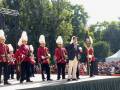 Una ceremonia militar con soldados en uniformes rojos y sombreros altos, un oficial en traje negro con corbata, y una bandera. La escena se realiza frente a un público en la calle con árboles y edificios al fondo.