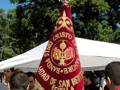 Banderín de la Banda de Música de San Benito de Sevilla, con detalles dorados y rojos, sobre un fondo blanco, en un ambiente festivo con personas al fondo y árboles en el cielo azul claro.