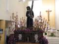 En una iglesia, un altar central con flores y velas encendidas. En el centro, una estatua de Jesús con un crucifijo en la mano izquierda. A los lados, candelabros de varios brazos y flores en macetas. En el fondo, una mesa con velas encendidas y un crucifijo en la pared.