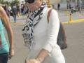 Una mujer con cabello largo y rubio, lleva gafas de sol negras y una bufanda estampada. Se dirige hacia la derecha, con un bolso de mano en el hombro izquierdo. En el fondo, se observa un área con varias personas y una motocicleta aparcada.