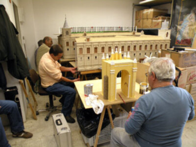 Un grupo de personas trabajando en un modelo de una fortaleza medieval, con detalles como torres y murallas. La imagen muestra a varias personas sentadas en sillas, uno de ellos está sentado sobre una caja. En el fondo se pueden ver estanterías con cartones y un mapa en la pared.