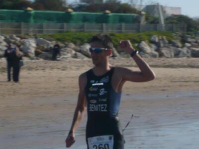 Un atleta en traje de baño negro con el nombre "BENITEZ" en la espalda, corriendo en una playa con rocas y un muro verde al fondo.