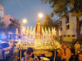 Fotografía nocturna de una procesión religiosa con una imagen de la Virgen en el centro, rodeada por velas y flores. La imagen está iluminada por farolas y luces de la calle, con edificios al fondo. Personas observan la procesión desde el borde de la calle.