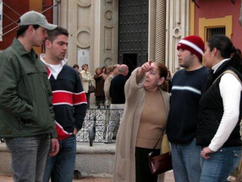 Una mujer con una bufanda beige y una chaqueta beige está señalando algo mientras camina con otros personas en un entorno urbano con una fuente y una estructura de piedra en el fondo.