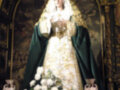 Virgen María en iglesia, vestida de blanco con capa verde y corona dorada. Fondo dorado con detalles artísticos. Base de flores blancas y velas en latón.