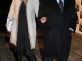 Una pareja camina juntos, la mujer con abrigo beige y pantalones negros, el hombre con traje negro y corbata. Ambos sonríen.