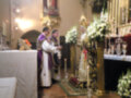 Imagen religiosa en un altar con dos sacerdotes, flores blancas y doradas, candelabros, vitrales y una imagen en el fondo.