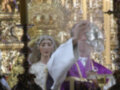 Imagen religiosa con un sacerdote vestido de blanco y morado, frente a una imagen de la Virgen María. El fondo muestra detalles dorados y cristalinos, probablemente parte de un altar o retablo.