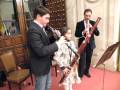 Tres personas, dos adultos y una niña, tocan instrumentos musicales en un ambiente formal. La imagen muestra a dos adultos tocando flautas y una niña tocando un instrumento de viento. La escena parece ser parte de una ceremonia o evento formal, con detalles como vestimenta elegante y un ambiente bien decorado.
