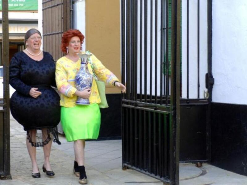 Dos mujeres caminan por una calle, una con un vestido negro y la otra con un vestido verde. La mujer del centro sostiene una botella de agua. Detrás de ellas, se puede ver un edificio con una puerta metálica y una ventana.