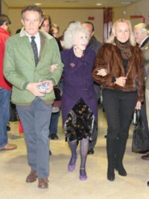 Una persona con abrigo verde y corbata, otra con vestido morado y tacones, y una tercera con abrigo marrón y botas altas. Todos están en un entorno interior, posiblemente una sala de conferencias o evento formal.