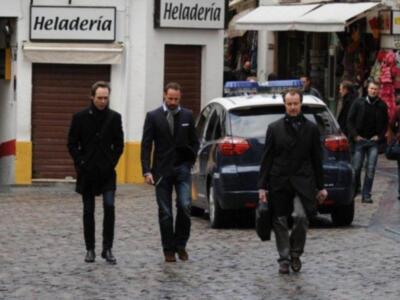 Tres hombres caminan por una calle adoquinada, uno de ellos con un bolso negro. En el fondo, se puede ver una heladería y otros negocios en un entorno urbano.