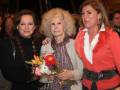 Tres mujeres posan juntas, una con un ramo de flores y otra con un abrigo rojo.