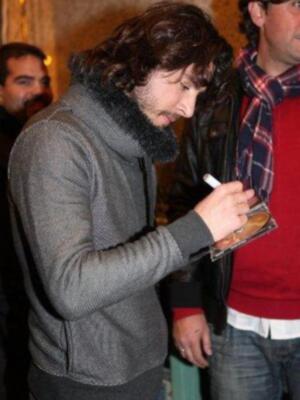 Un hombre con una chaqueta gris y gorro de lana está firmando un autógrafo en un papel. Detrás de él, otro hombre con una chaqueta roja y bufanda de lana.
