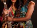 Dos mujeres posan con una guitarra acústica, vestidas con vestidos de colores vibrantes.