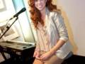 Una mujer con cabello rojo largo y una blusa de terciopelo beige posa frente a un teclado musical.