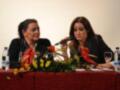 Conferencia con dos mujeres hablando en un escenario con micrófono y flores en la mesa.
