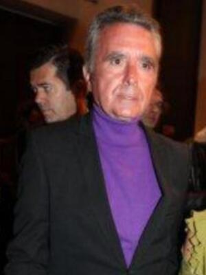 Un hombre con cabello corto y canoso, lleva una chaqueta negra y un suéter violeta. La imagen parece ser de una celebración o evento formal, con otros asistentes en el fondo.