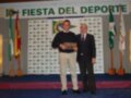 Fiesta del Deporte, con dos hombres en trajes posando junto a un trofeo. Banderas y decoración de fondo.