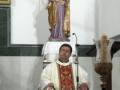 Un sacerdote vestido con un hábito blanco y rojo, posa frente a una estatua de la Virgen María con el Niño Jesús en un altar. La imagen está en un interior, posiblemente una iglesia, con detalles como la estatua y el hábito del sacerdote.