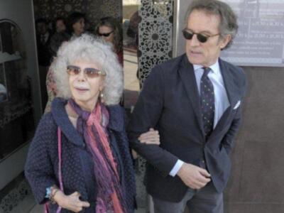 Dos personas, una mujer mayor con gafas y un hombre con traje y corbata, caminan juntos.