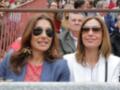 Dos mujeres con gafas de sol sonríen mientras observan una competición deportiva. La imagen muestra a varias personas en el fondo, probablemente espectadores o participantes de la actividad.