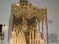 Un altar elaborado con velas altas y flores blancas, rodeado por una gran mantilla dorada. En el centro se encuentra un personaje vestido con trajes tradicionales, rodeado de luces y adornos. La atmósfera es festiva y ceremonial.