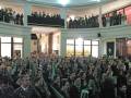 Una multitud en un auditorio, muchos con banderas verdes y uniformes militares.