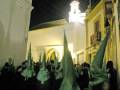Una procesión nocturna en una ciudad con edificios blancos y amarillos, donde los participantes llevan capuchas verdes. La imagen muestra una escena de la Semana Santa en España, con un cielo oscuro y luces brillantes.