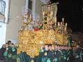 Una procesión religiosa con una imagen grande y dorada, vestidos de negro y verde, frente a un edificio histórico en la noche.