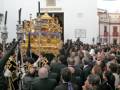 Una procesión religiosa con una imagen dorada en el centro, rodeada por una multitud de personas vestidas con trajes formales, frente a una iglesia con una fachada blanca y detalles dorados.