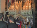 Una procesión religiosa con una imagen de la Virgen en el centro, rodeada por velas encendidas y flores blancas. Personas vestidas formalmente observan la escena, con un altar elaborado en el fondo.