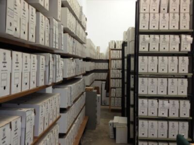Un gran almacén lleno de estanterías repletas de cajas blancas identificadas con números y letras. La iluminación es suave, creando sombras en las estanterías de madera. Los cajas parecen estar organizadas por categorías, posiblemente para un archivo o biblioteca.