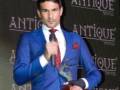 Un hombre en traje azul con corbata roja y un corte de pelo corto, posando en una alfombra roja con el logo "Antique" en el fondo.
