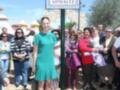 Una mujer en un vestido verde se encuentra sobre una placa de calle con el nombre "Avenida Eva González". La imagen muestra a varias personas observando la escena.