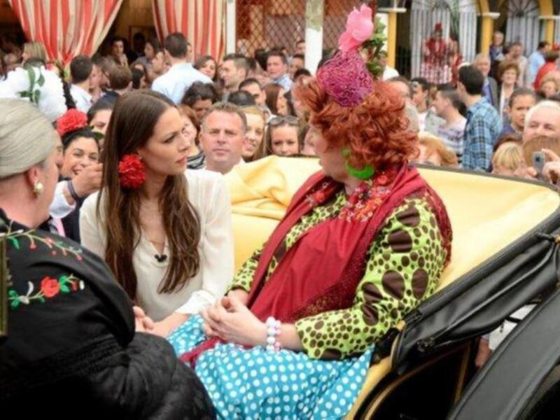 Dos personas vestidas con trajes tradicionales se sientan en un coche, rodeadas por una multitud de personas con trajes y sombreros. La escena parece ser parte de una celebración festiva o tradicional, posiblemente en España.