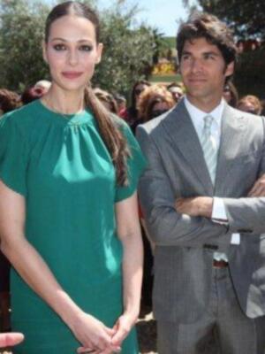 Una mujer con vestido verde y un hombre en traje gris están posando para la foto. El fondo muestra a varias personas, algunos con sombrillas y otros con cabello largo.