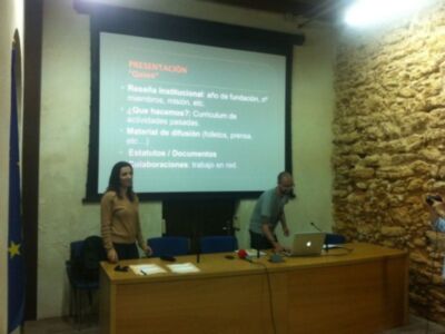 Presentación institucional en una sala de conferencias. Una mujer presenta ante un grupo, con un proyector mostrando información sobre la institución y actividades pasadas. Un hombre está en el fondo, con un dispositivo de grabación. La imagen incluye una mesa con documentos y un portátil, junto a una bandera europea.