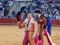 Un torero en traje tradicional sostiene un sombrero mientras camina por el ruedo de la plaza de toros. La multitud observa desde los palcos detrás del ruedo.