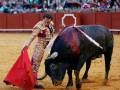 Un torero con un traje tradicional de color dorado y rojo, sostiene una capa roja mientras interactúa con un toro en el centro de la imagen. La arena es de tierra y hay una pared roja al fondo con un logo blanco en la parte superior. La audiencia está sentada en gradas detrás de la pared.