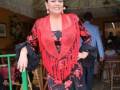 Una mujer vestida con un traje flamenco tradicional, posando en una fiesta. La imagen muestra una decoración típica de eventos culturales, con flores y sillas en el fondo.