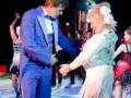 Un hombre y una mujer están bailando en un escenario de circo. El hombre lleva un traje azul y la mujer una falda larga con flecos. En el fondo, se pueden ver otros bailarines y la iluminación característica del circo.