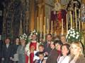 Una procesión religiosa en una iglesia con personas vestidas de gala, un altar dorado y flores blancas.