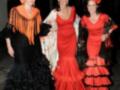 Tres mujeres vestidas con trajes de flamenca, con colores rojo y negro, posando para la cámara en una calle iluminada por faroles.